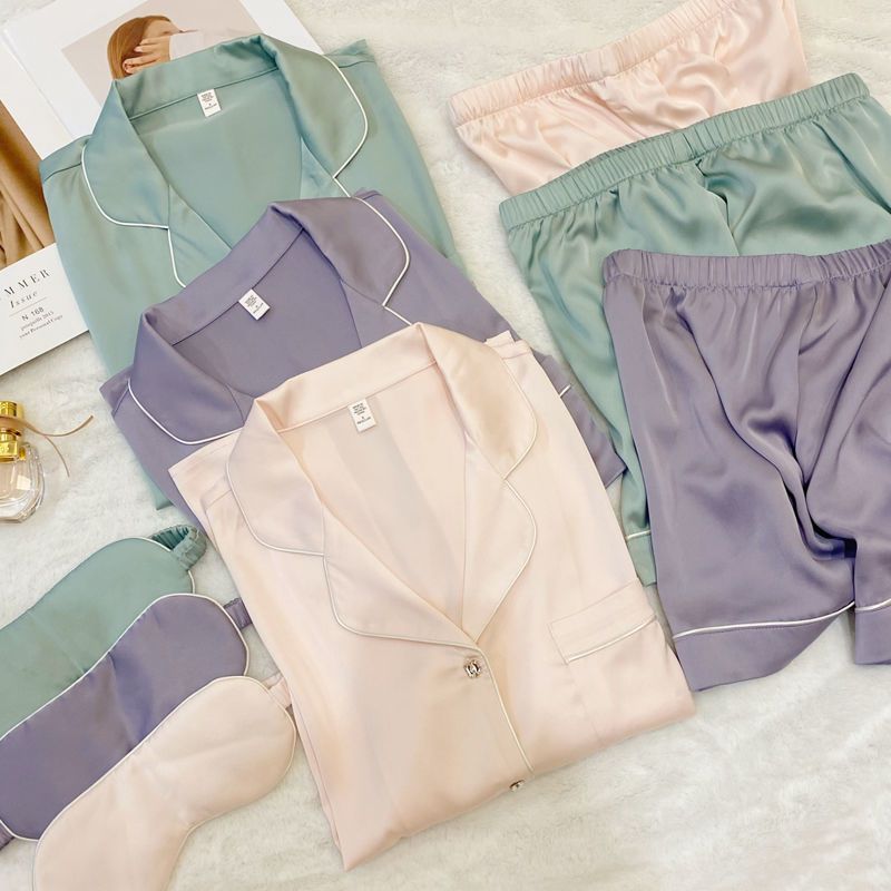 Three satin pajama sets in soft pastel colors are arranged on a white textured blanket, including matching sleep shirts and shorts in mint green, lavender, and blush pink, with a matching sleep mask and a small perfume bottle nearby.
