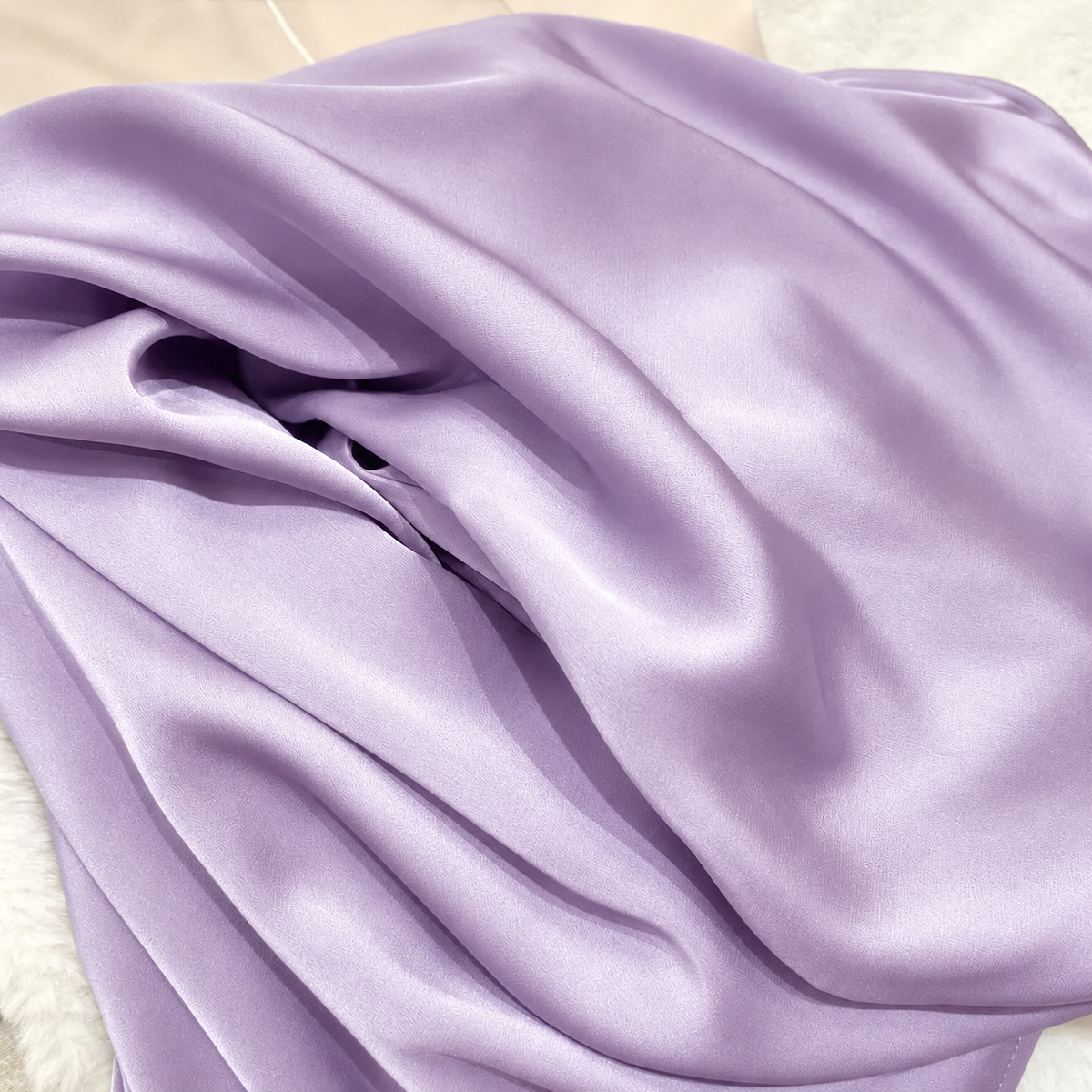 A close-up of draped lavender satin fabric with soft folds and a subtle sheen, resting on a light neutral surface.