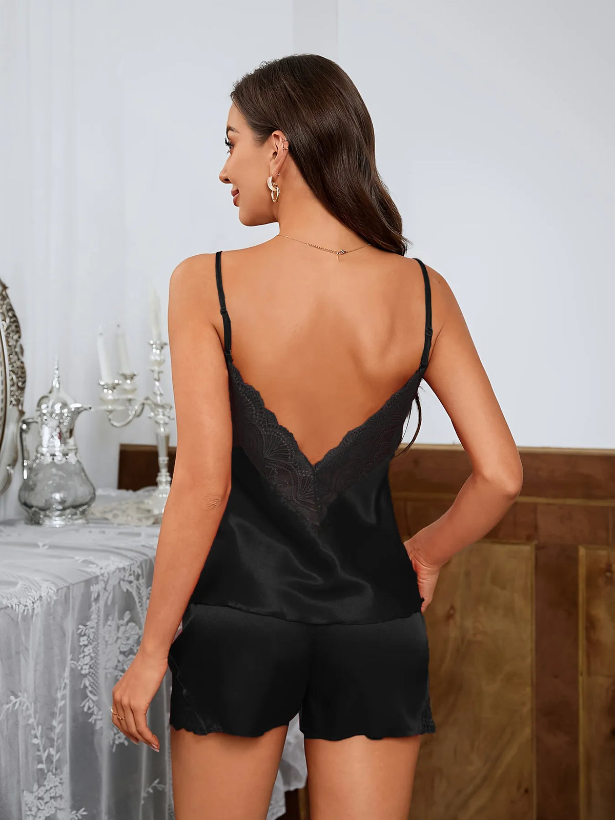 A woman shown from the back wearing a black satin camisole with a lace-trimmed open back and matching black satin shorts, posed indoors against a light wall with furniture and decor in the background.