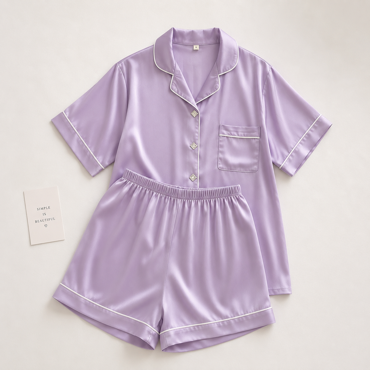 A lavender purple two-piece pajama set with white piping, consisting of a short-sleeve button-front top with a collar and a matching shorts set, laid flat on a light beige background.