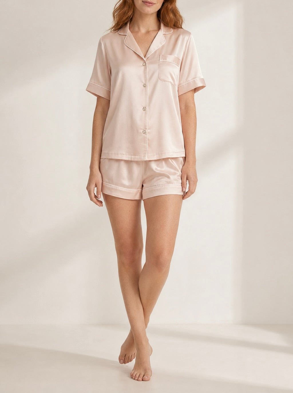 A woman modeling a light blush pink two-piece pajama set with a short-sleeve button-front shirt and matching shorts, photographed in a softly lit studio setting.